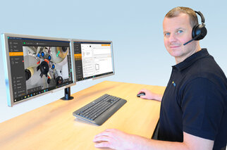A man is sitting at a desk with two monitors displaying technical software and a camera view. He is wearing a headset.