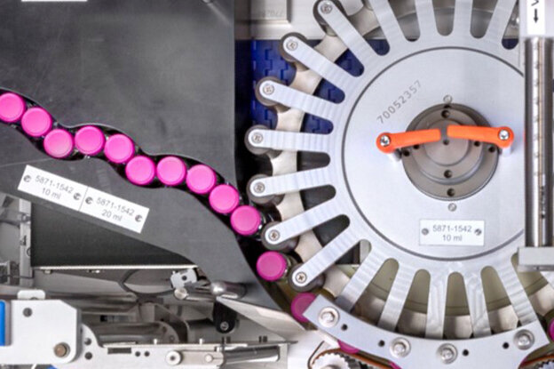 A person in protective gear is holding a syringe filled with a liquid, while a technical machine with colorful components is visible in the background.