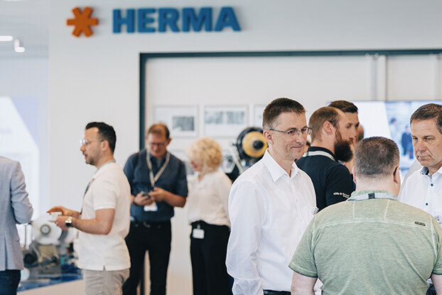 People in formal attire are standing in a modern space, discussing and interacting, with a HERMA sign in the background.