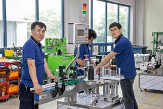 Three workers in blue shirts operate a machinery setup in a factory, surrounded by tools and equipment.