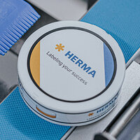 Round container with the print 'HERMA' and the slogan 'Labeling your success,' surrounded by blue and white elements.