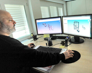 A man is working at a computer with two monitors displaying 3D models. The desk is equipped with office supplies.