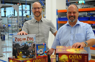 Two men are standing in a toy store showcasing board games, including 'Ticket to Ride: Europe,' 'Halli Galli,' and 'Catan Plus.