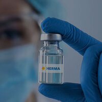 A person wearing blue gloves is holding a small glass bottle labeled 'HERMA'.