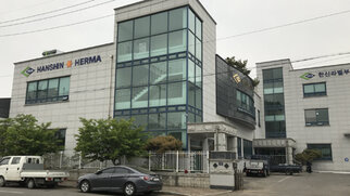 Modern office buildings with large windows, labeled 'HANSHIN' and 'HERMA'. Surrounded by trees and parked cars.