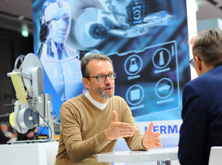A man is talking to another man while a technical presentation and a robot are visible in the background.