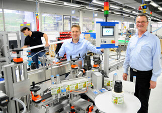 Two men are standing at a production line in a factory, while a third employee is working in the background.