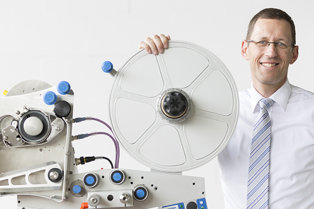 A man in a shirt and tie is holding a large disc and standing next to technical equipment with hoses and connections.