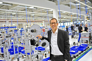 A man in a suit stands in a modern production hall, holding a device. In the background, workstations and blue containers are visible.