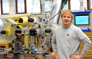 A man stands next to a modern machinery setup for processing materials, surrounded by technical equipment and screens.