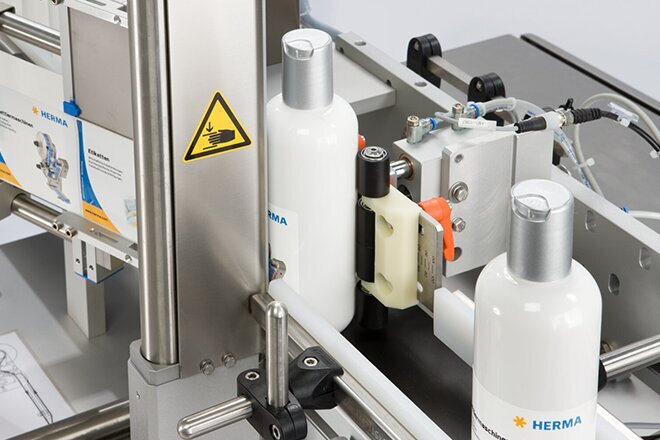 A bottling machine with white bottles on a conveyor belt, equipped with various technical components and warning signs.