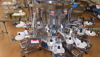 Automatic labeling machine with multiple stations that applies labels to products. Control panel visible in the foreground.