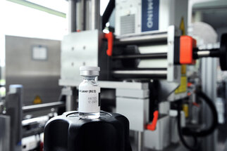 A vaccine vial is standing on a black lid, with machines for vaccine production visible in the background.