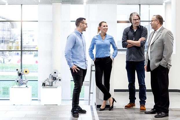 Four people are talking to each other in a modern office, while in the background, an industrial building and a sprawling facility are visible.