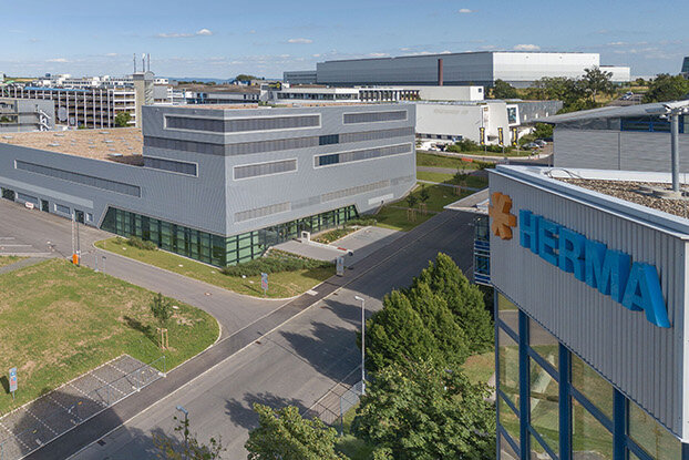 Four people are talking to each other in a modern office, while in the background, an industrial building and a sprawling facility are visible.