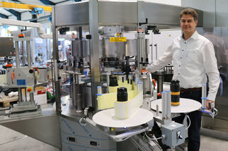 A man stands next to a modern bottling and labeling machine in a production hall, surrounded by bottles and machinery.
