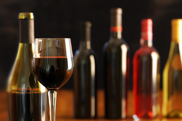 A glass of red wine stands in front of several wine bottles in different colors on a table.