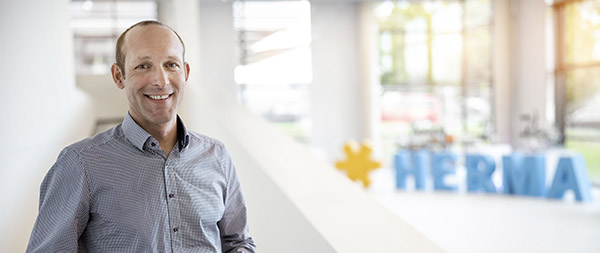 A man in a checkered shirt smiles in a modern office with the sign "HERMA" in the background.
