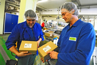 Two men in blue work uniforms and hairnets are holding boxes in a production environment and checking their contents.